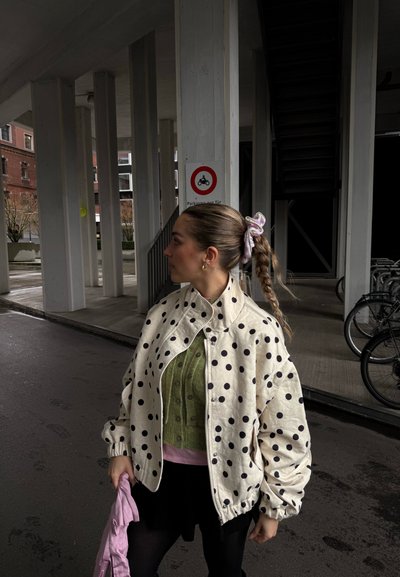 Mujer con coleta trenzada que lleva una chaqueta de lunares y un suéter verde, de pie bajo una estructura de estacionamiento cerca de bicicletas aparcadas.