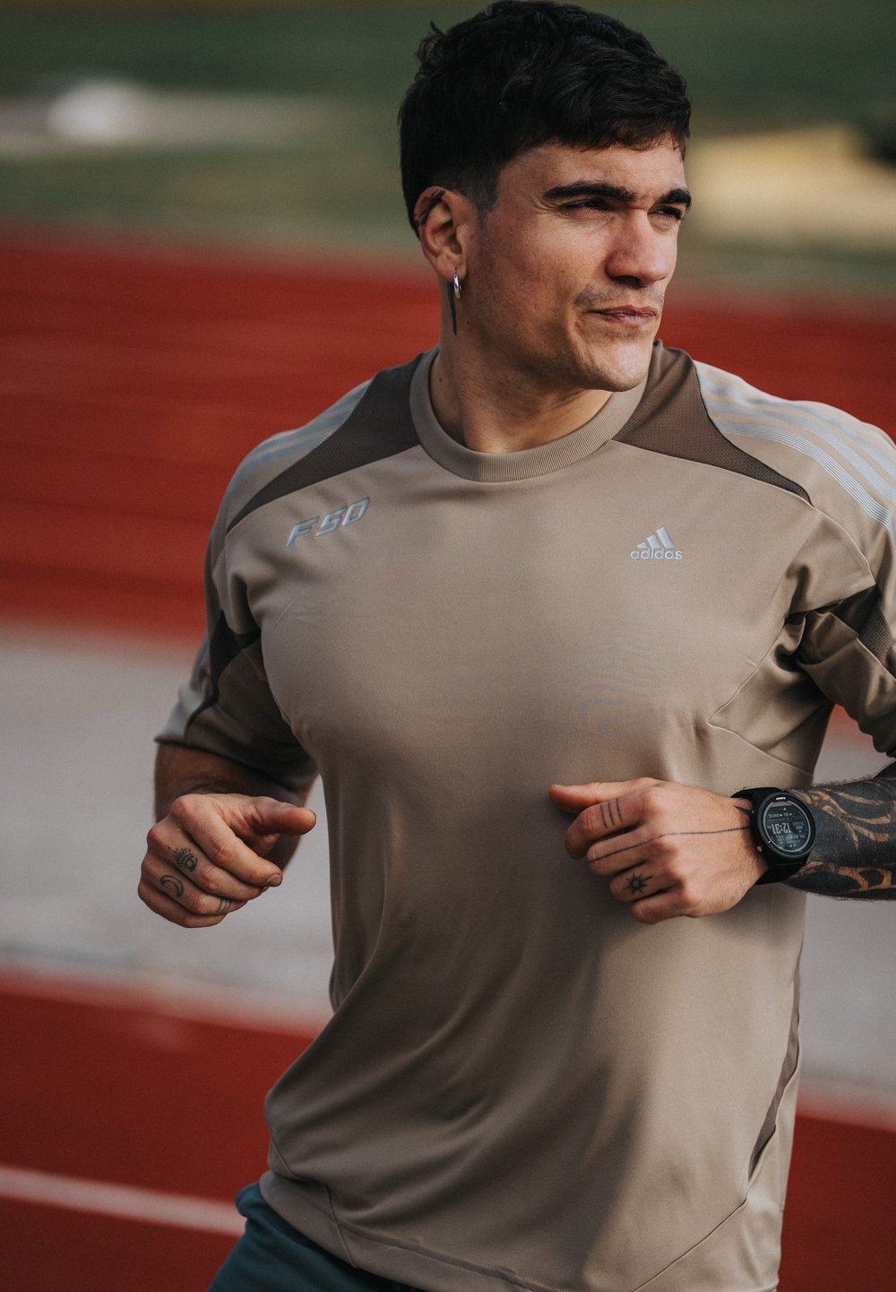 T-shirt de sport à manches courtes beige avec des accents foncés sur les épaules, arborant un logo bleu clair; montre au poignet; fond de piste de course rouge.