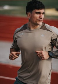 T-shirt de sport à manches courtes beige avec des accents foncés sur les épaules, arborant un logo bleu clair; montre au poignet; fond de piste de course rouge.