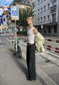 Jeune personne aux cheveux blonds portant un débardeur blanc et un pantalon large noir, drapant une veste en fourrure beige sur un bras dans une rue urbaine.