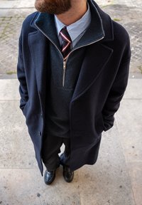 Casaco de lã azul-marinho sobre um suéter escuro, combinado com uma camisa de colarinho azul claro e uma gravata riscada; sapatos sociais pretos completam o visual.