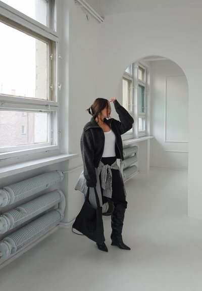 Mujer con chaqueta negra, blusa blanca y botas, de pie en una habitación luminosa con grandes ventanas y una puerta arqueada, sosteniendo un bolso negro texturizado.