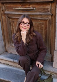 Jeune femme en tenue marron et lunettes, assise sur des marches en pierre, tenant un sac à main vert, souriant et reposant son menton sur sa main.