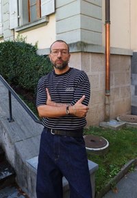 Mężczyzna w okularach i kolczykach stoi z założonymi rękami, nosząc koszulę w paski i ciemne dżinsy w miejskim otoczeniu na świeżym powietrzu.