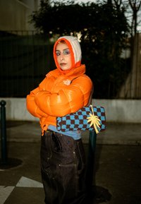 Personne portant une doudoune orange et une capuche en tricot tenant un sac à carreaux bleu et marron avec un détail de palmier jaune.