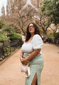 Blusa branca de mangas bufantes com tecido texturizado, combinada com uma saia verde com padrão floral e uma fenda alta na coxa. Segura uma pequena bolsa bege.