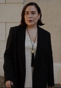 Blazer nero doppiopetto su un vestito leggero color crema, con una lunga collana che presenta una perla rotonda e un dettaglio pon pon.
