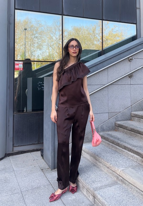 Femme aux longs cheveux noirs et lunettes se tient près d'escaliers en pierre, portant un haut marron à une épaule, un pantalon assorti, des ballerines roses et tenant un sac rose.