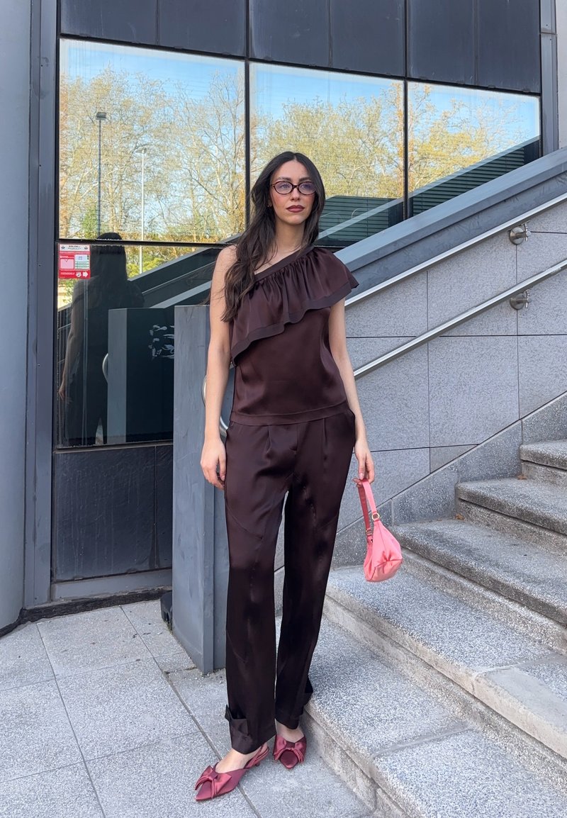 Femme aux longs cheveux noirs et lunettes se tient près d'escaliers en pierre, portant un haut marron à une épaule, un pantalon assorti, des ballerines roses et tenant un sac rose.