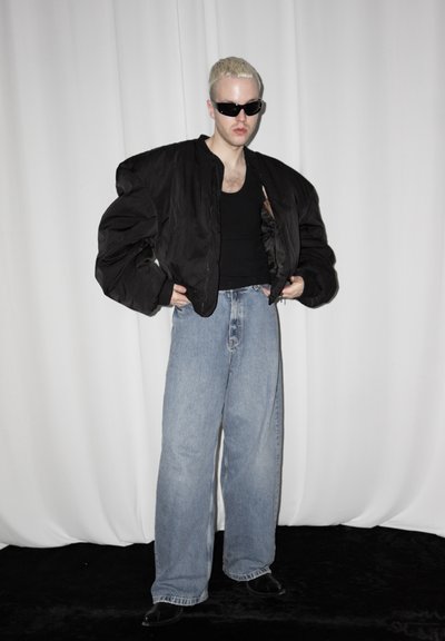 Persona con cabello rubio corto que lleva gafas de sol negras, chaqueta negra extragrande, camiseta negra, vaqueros azules de pierna ancha y zapatos negros, de pie frente a una cortina blanca.