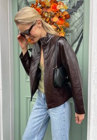 Brown leather jacket with a zip front, high collar, and a relaxed fit, teamed with light blue denim jeans and a small black handbag.