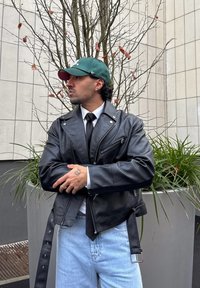 Homme portant une casquette de base-ball verte, une veste en cuir noire, une chemise blanche, une cravate noire et un jean bleu clair, debout les bras croisés près de plantes en pot et d'un arbre dépourvu de feuilles.