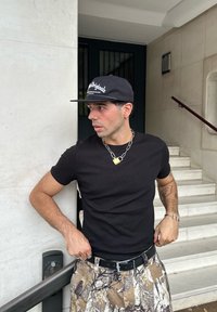 T-shirt preto ajustado, calças de camuflagem com padrão, e um boné preto com texto branco. Colar de corrente prateada com um cadeado amarelo.