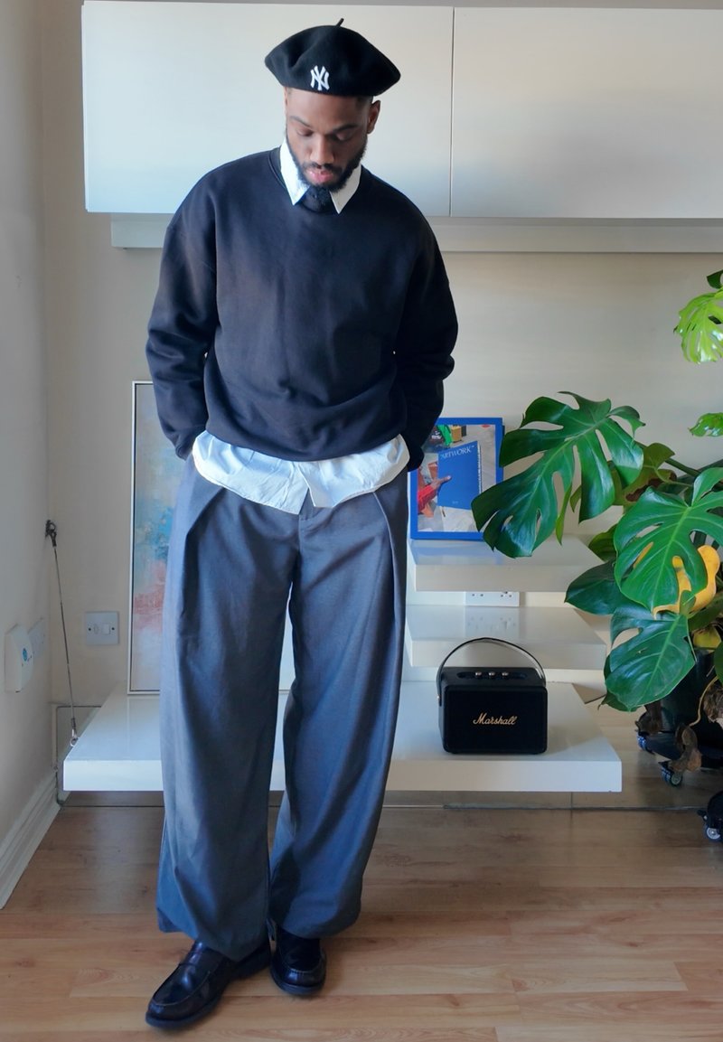 Black sweatshirt, white collared shirt underneath, gray wide-leg trousers, black beret with logo, black shoes, indoor plant in background.