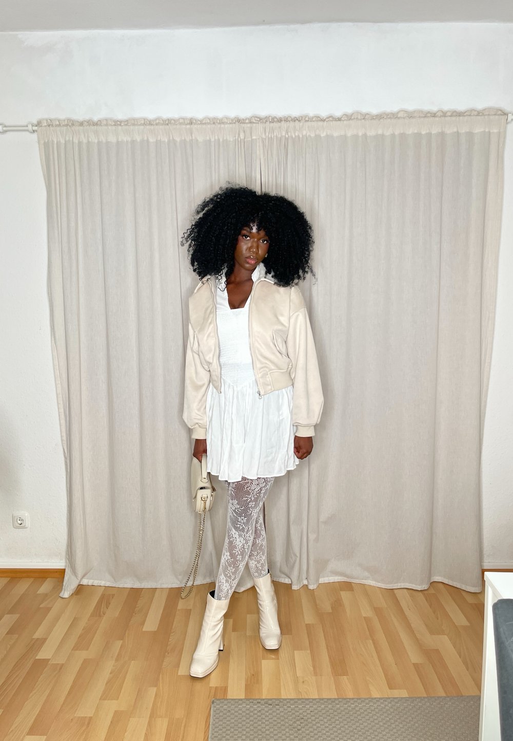 Beige bomberjack over een witte jurk, gecombineerd met witte kanten tights en witte enkellaarsjes. Kort haar gestyled in volumineuze krullen.