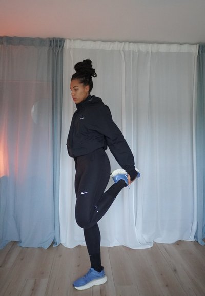 Chaqueta negra recortada, mallas negras, zapatillas deportivas azules; pose de estiramiento. Habitación con cortinas claras y suelo de madera.