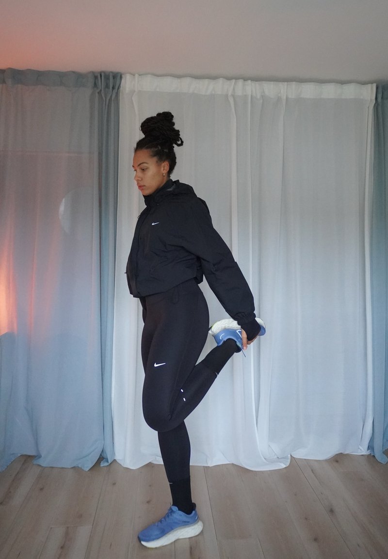 Casaco preto cropped, leggings pretas, sapatilhas desportivas azuis; pose de alongamento. Quarto com cortinas claras e chão de madeira.