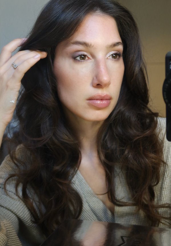 Vrouw met lang golvend donker haar, die een beige trui draagt, haar haar aanraakt en iets naar de zijkant kijkt met een neutrale uitdrukking.