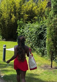 Piel bronceada y cabello largo y ondulado. Vestido rojo con lunares blancos, sosteniendo un bolso blanco con detalles rojos, caminando sobre hierba verde cerca de follaje.