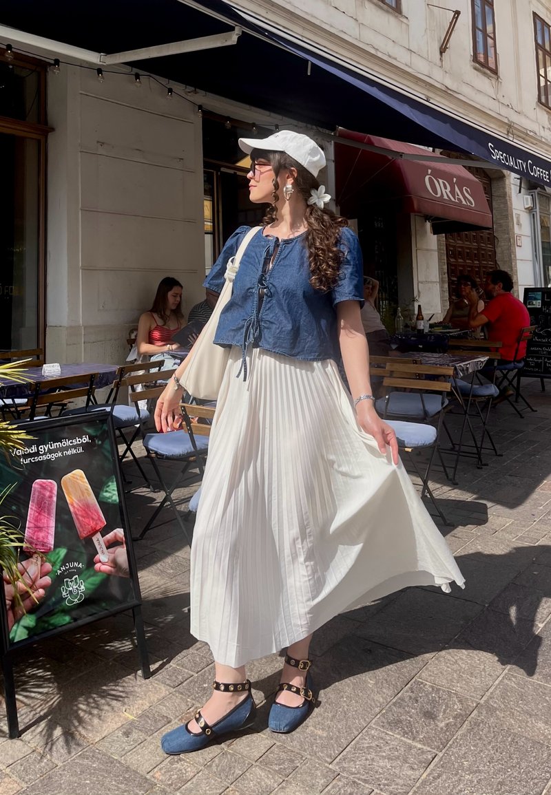Femme en jupe blanche, haut bleu, casquette blanche et chaussures bleues debout devant un café avec terrasse extérieure et des personnes en train de dîner.