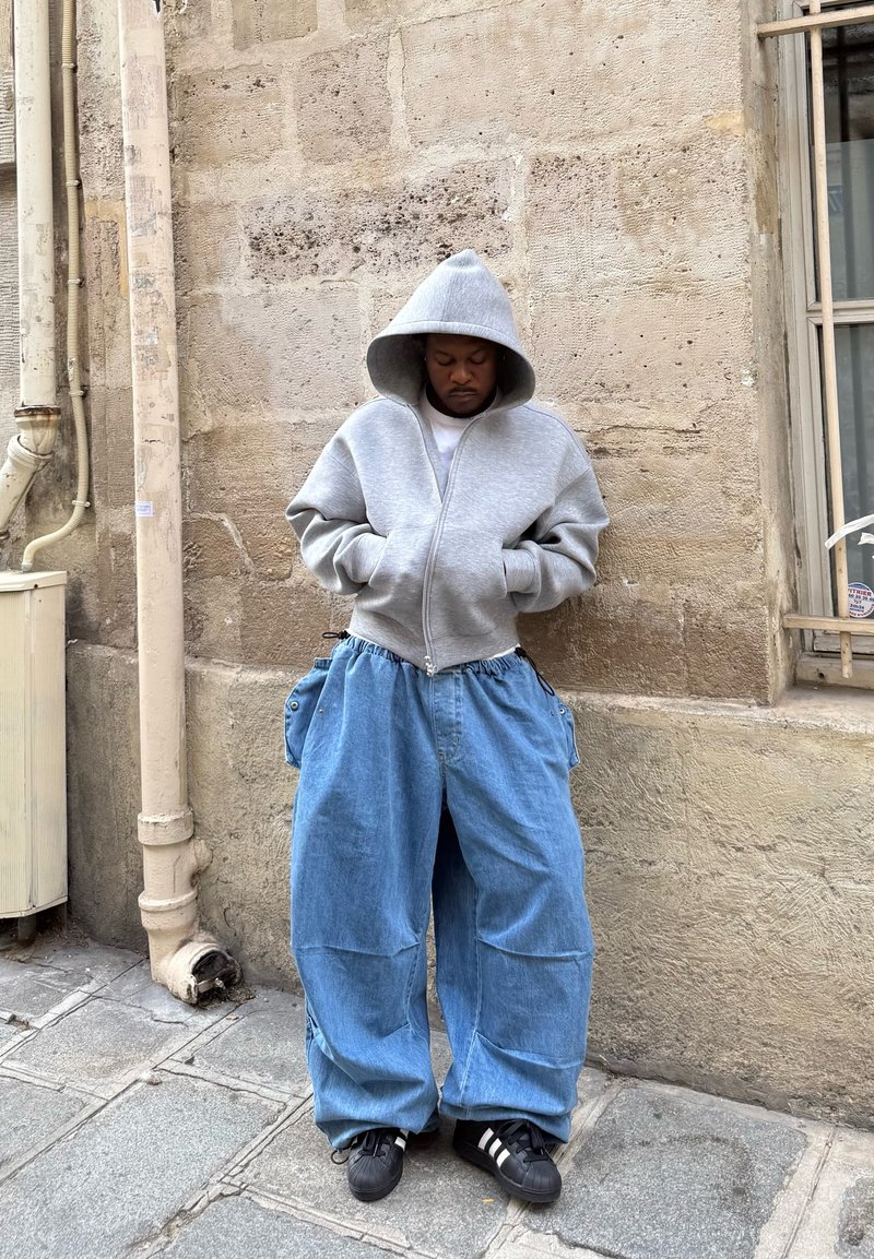 Haut à capuche gris zippé avec une capuche et des poches avant, associé à un pantalon baggy en denim bleu. Baskets noires avec des rayures blanches. Fond de mur en pierre clair.
