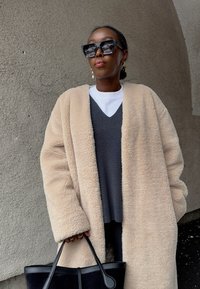 Άτομο που φοράει μεγάλα μαύρα γυαλιά ηλίου, μπεζ χνουδωτό παλτό, γκρι πουλόβερ πάνω από λευκό πουκάμισο, κρατώντας μαύρη τσάντα, στέκεται μπροστά σε υφή τοίχου.