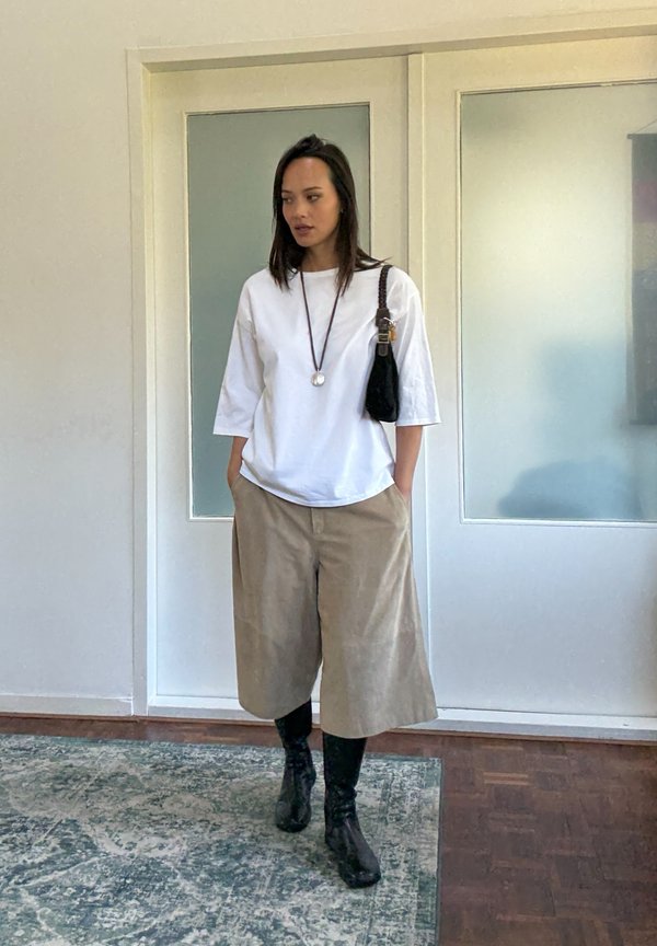 Camicia bianca a maniche lunghe, pantaloni beige a gamba larga, borsa nera a mano e stivali neri al ginocchio. Collana con un pendente rotondo come accento.