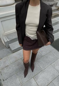 Blazer de rayas negras sobre una camiseta beige con encaje, combinado con una falda corta marrón oscuro con tachuelas y botas marrones altas hasta la rodilla.