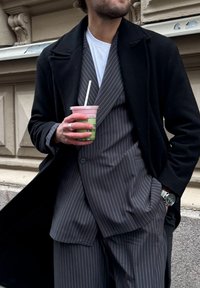 Fato cinza riscado com uma camisa branca e casaco preto. Segurando uma chávena com uma bebida rosa e verde. Relógio prateado no pulso, de design minimalista.