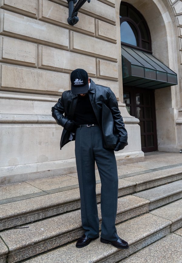 Férfi fekete bőrdzsekiben, fekete sapkában, kesztyűben és sötétszürke nadrágban, aki kőlépcsőn áll egy klasszikus épület előtt, amelynek boltíves ajtaja van.