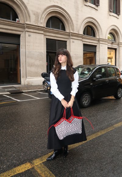Vestido negro con una blusa blanca, sosteniendo un bolso de mimbre con un patrón de zigzag en blanco y negro y asas rojas. Pavimento mojado y coches al fondo.