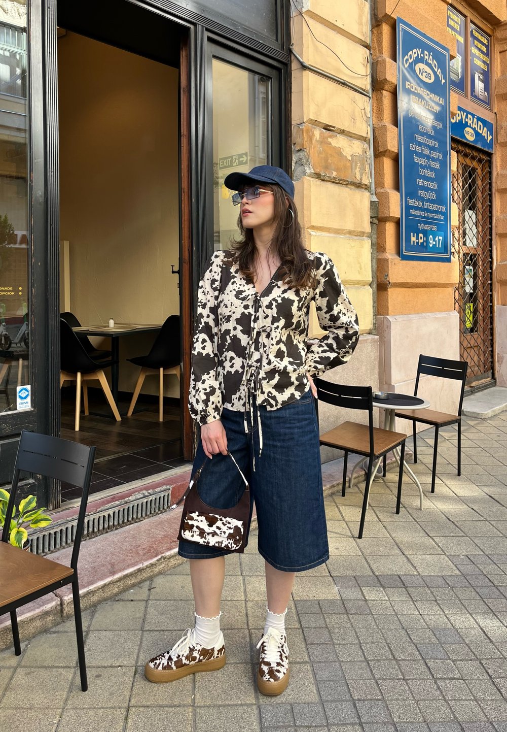 Ilga rankovė blouse su rudos ir grietinėlės spalvos karvės raštu, derinama su džinsinėmis kelnių knygelėmis, taškuotais sportbačiais ir derančia rankine, visiškai atsitiktinėje aprangoje.