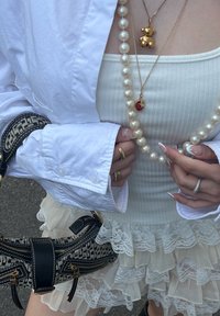 Camisa branca com botões, blusa canelada creme, saia de renda em camadas, colar de urso de pelúcia dourado, pérola e pendente vermelho, bolsa preta estampada com zíperes.