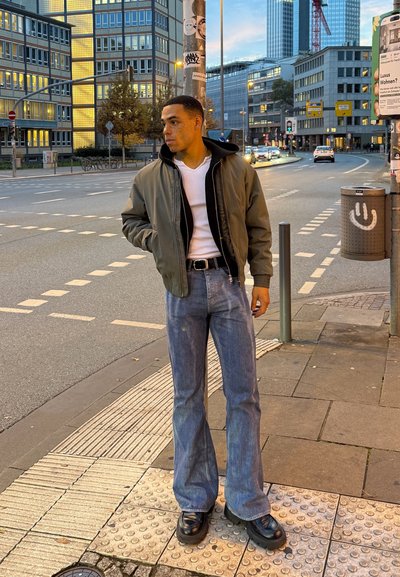Joven con chaqueta verde, camisa blanca, jeans y zapatos negros, de pie en una acera de la ciudad cerca de un paso de peatones al anochecer.