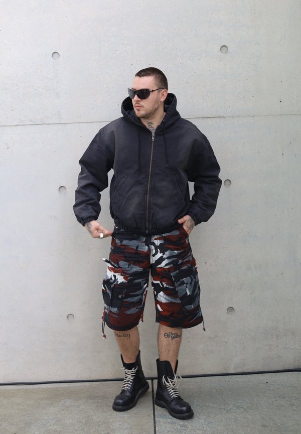 Svart hettejakke med glidelås, grafiske kamuflasje cargo shorts i rød, grå og hvit, svarte militærstøvler med snøring, står mot en betongvegg.