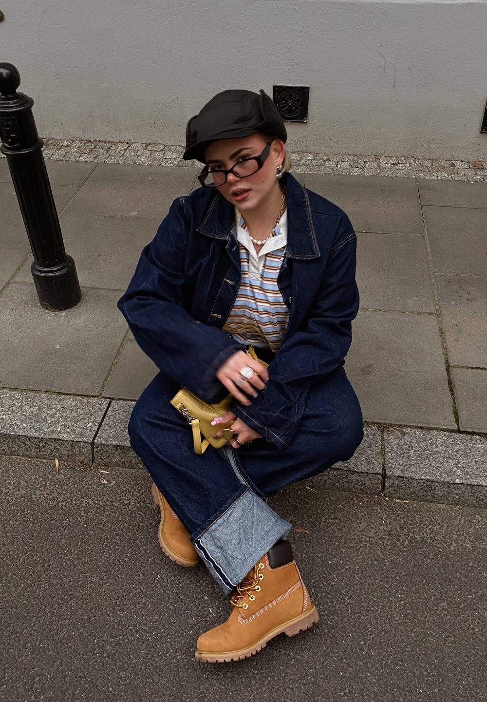 Veste en denim et jeans à jambes larges avec des poignets bleu clair contrastants, bottes de travail beiges, casquette noire et sac bandoulière jaune.