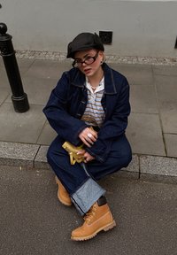 Veste en denim et jeans à jambes larges avec des poignets bleu clair contrastants, bottes de travail beiges, casquette noire et sac bandoulière jaune.