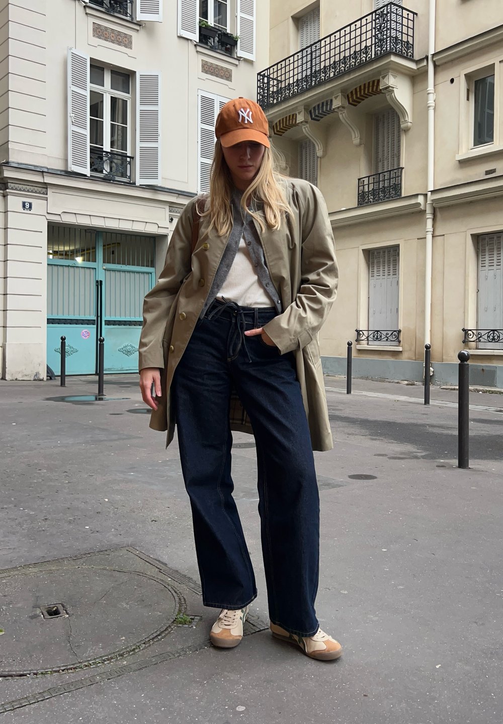 Giovane che indossa un trench beige, jeans larghi scuri, sneakers bianche e un cappello arancione, in piedi su una strada cittadina con edifici sullo sfondo.