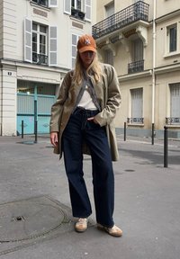 Giovane che indossa un trench beige, jeans larghi scuri, sneakers bianche e un cappello arancione, in piedi su una strada cittadina con edifici sullo sfondo.