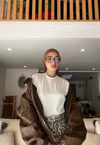 Hnedá kožená oversized bunda cez biely top, doplnená o nohavice s leopardím vzorom a strieborným opaskom. Hnedý šatka na hlave a veľké okuliare dotvárajú vzhľad.