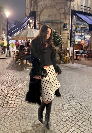 Abrigo negro de piel sintética sobre una camiseta negra, falda asimétrica blanca con lunares negros y botas negras hasta la rodilla. Caminando por una calle adoquinada de noche.