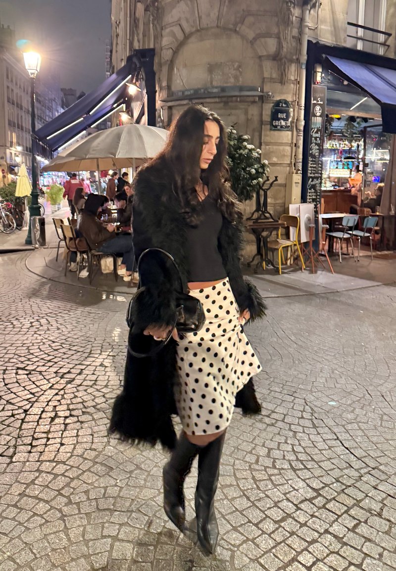 Cappotto in pelliccia sintetica nera sopra una maglietta nera, gonna asimmetrica bianca con pois neri e stivali neri fino al ginocchio. Camminando su una strada lastricata di ciottoli di notte.