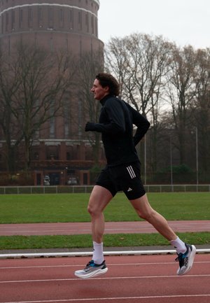 Hombre con ropa deportiva negra corriendo en una pista al aire libre, con un gran edificio de ladrillo y árboles sin hojas al fondo.