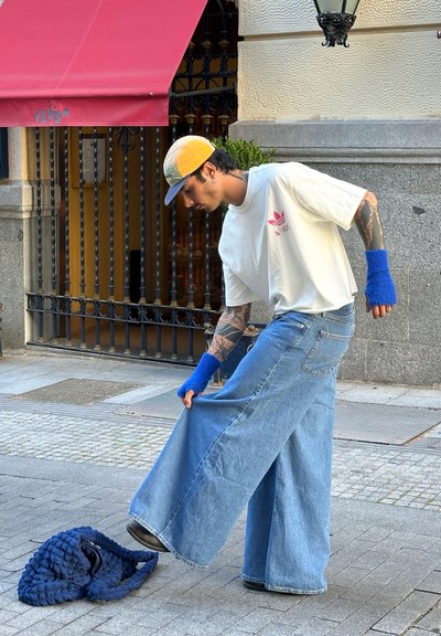Pantalones de mezclilla de pierna ancha en azul claro con una textura desgastada, combinados con una camiseta blanca que lleva un logo rosa y guantes sin dedos azules.
