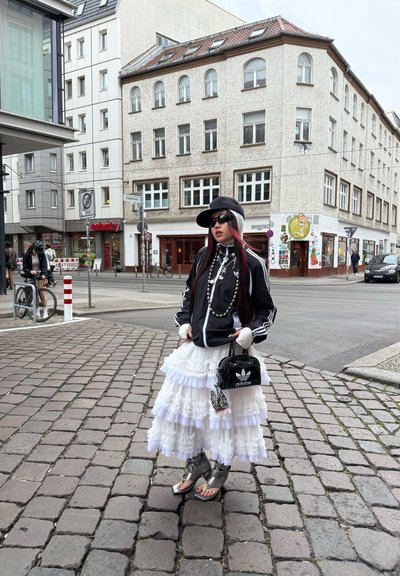 Chaqueta negra con cremallera y rayas blancas, falda blanca con volantes en capas, sandalias de plataforma plateadas, collar de perlas, bolso negro. Entorno urbano.