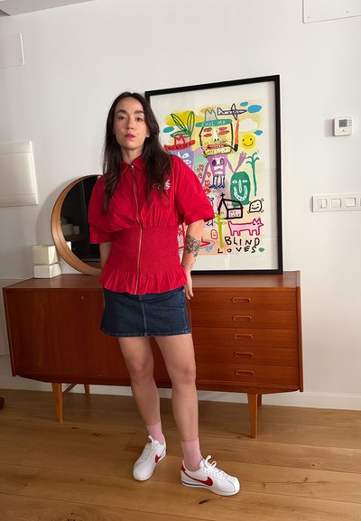 Mujer con blusa roja, falda vaquera y zapatillas blancas está delante de un aparador de madera con una obra de arte abstracta y colorida en pared blanca.