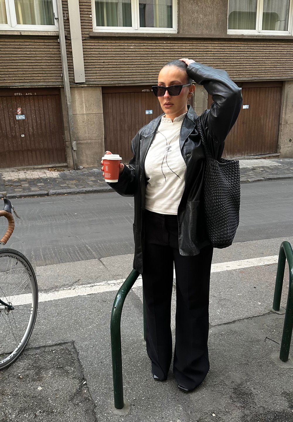 Femme en veste en cuir noire et lunettes de soleil debout sur le trottoir tenant une tasse de café, portant un sac fourre-tout noir tissé, à côté d'un vélo garé.