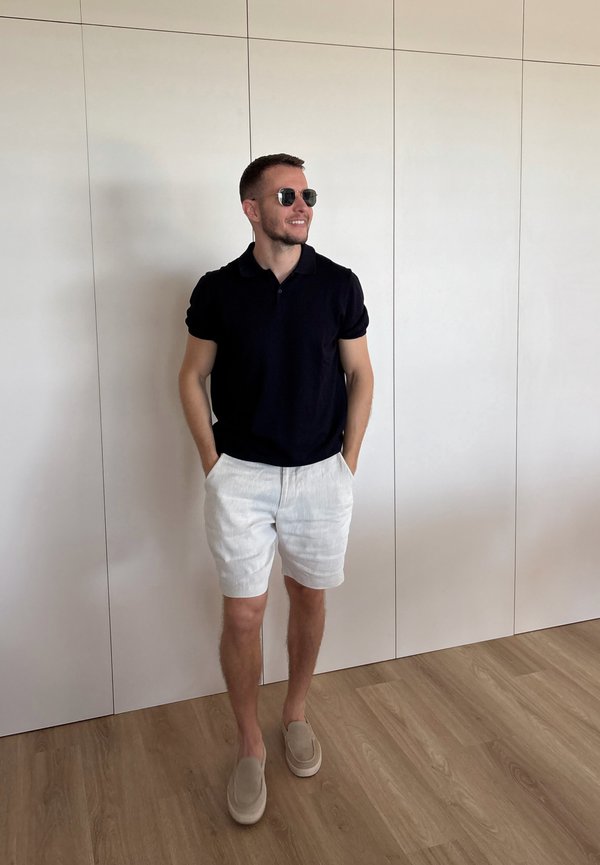 Jonge man met een zwarte polo, witte korte broek, beige instapschoenen en een zonnebril, staand met handen in de zakken tegen een effen witte muur.