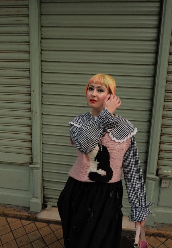Suéter rosa de punto con diseño de un gato negro, combinado con una falda negra y una camisa a cuadros con un cuello de encaje blanco; sostiene una bolsa rosa.