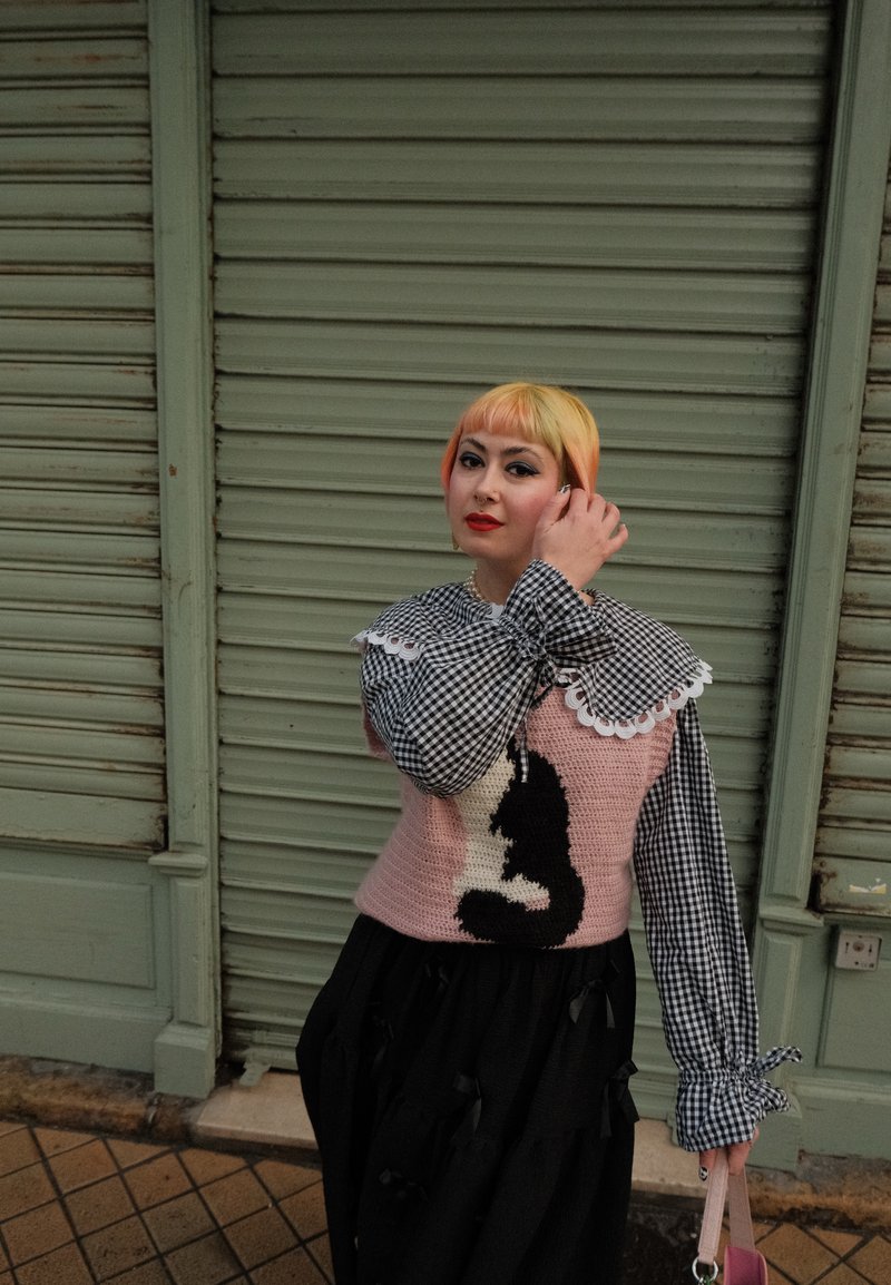 Pull rose tricoté présentant un motif de chat noir, associé à une jupe noire et une chemise à carreaux avec un col en dentelle blanche ; tenant un sac rose.